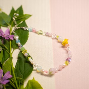 May include: A colorful beaded necklace with a yellow duck charm. The necklace is made with various pastel-colored beads, including clear, pink, green, and blue. The necklace is lying on a white and pink surface with green leaves and pink flowers.