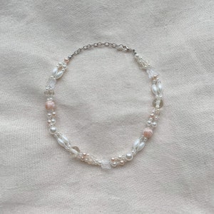 Peut inclure: Un collier d&eacute;licat en perles blanches et roses avec un fermoir argent&eacute;. Le collier pr&eacute;sente une vari&eacute;t&eacute; de perles, notamment des perles, des perles de verre transparentes et de petits pendentifs en forme de papillon.