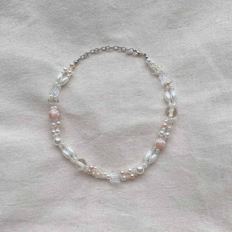 Peut inclure: Un collier d&eacute;licat en perles blanches et roses avec un fermoir argent&eacute;. Le collier pr&eacute;sente une vari&eacute;t&eacute; de perles, notamment des perles, des perles de verre transparentes et de petits pendentifs en forme de papillon.