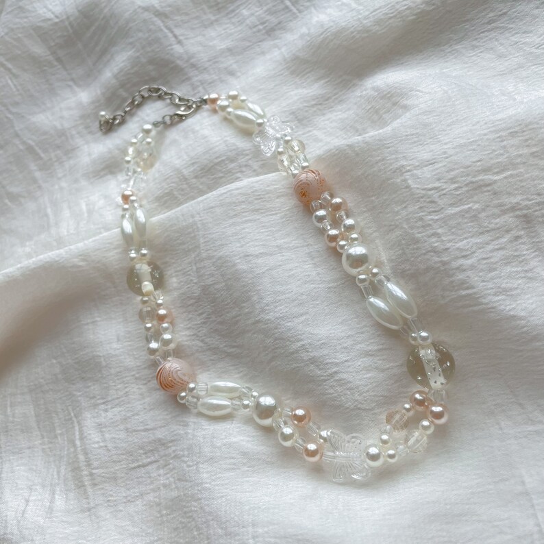 Peut inclure: Un collier d&eacute;licat &agrave; double rang de perles blanches et roses, comprenant des perles nacr&eacute;es et des breloques papillon transparentes.