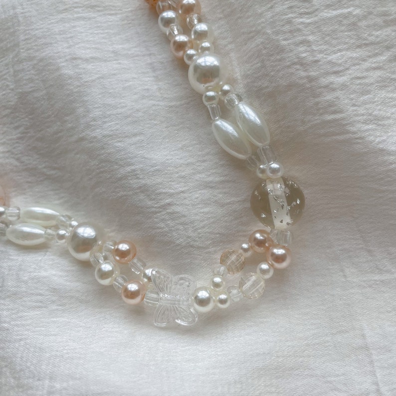 Peut inclure: Un collier &agrave; double rang de perles blanches et p&ecirc;che, avec un pendentif papillon transparent et une perle transparente avec des paillettes argent&eacute;es.