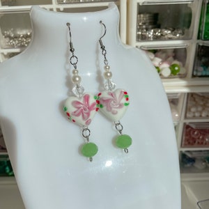 Pode incluir: Um par de brincos de prata com contas de vidro em forma de coração branco. As contas têm um design floral rosa e vermelho. Os brincos também têm uma conta de vidro verde pendurada em cada coração.