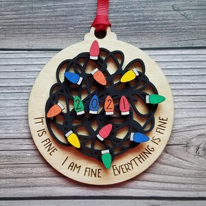 May include: A wooden Christmas ornament shaped like a lightbulb with a tangled string of lights. The ornament is decorated with colorful lights and the text "It is fine I am fine Everything is fine" and the year "2021".