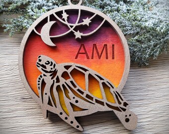 May include: A circular wooden ornament featuring a laser-cut sea turtle silhouette layered over a gradient background transitioning from deep purple to orange and yellow. The design includes a crescent moon and stars, with the name AMI engraved.