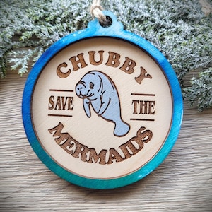 May include: A round wooden ornament with a blue-stained edge and a natural wood center. It features a laser-engraved illustration of a manatee in the middle, surrounded by the text CHUBBY SAVE THE MERMAIDS, with a jute twine loop for hanging.