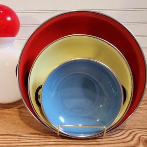 May include: A white and red bowling pin-shaped decorative object next to a set of three nested bowls. The bowls are red, yellow, and blue, with silver rims. The bowls are held in a gold-colored metal stand.