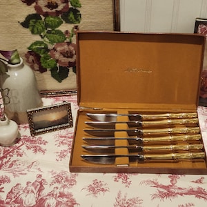 May include: A set of six stainless steel knives with gold-colored bamboo-style handles, presented in a brown box with a tan interior. The knives are arranged neatly, suggesting a dining or serving set.