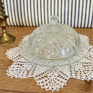 May include: A clear glass butter dish with a domed lid, resting on a white lace doily. The dish has a decorative, cut-glass pattern. A brass candlestick is visible in the background, set against a striped fabric.