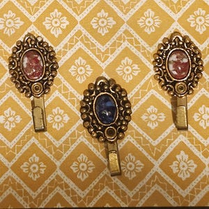 May include: Three metal wall hooks with an antique brass finish, each featuring an oval-shaped decorative head. Two hooks contain a mottled red and white stone inlay, while the central hook features a solid dark blue stone inlay.