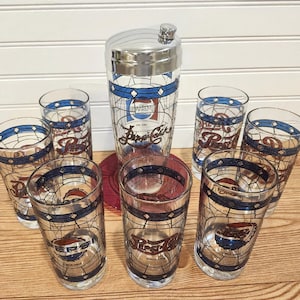 May include: A vintage Pepsi-Cola cocktail set featuring a chrome-topped shaker and six matching highball glasses. The glasses and shaker have a stained-glass design with blue and red accents, and the Pepsi logo. The set is displayed on a wooden surface.