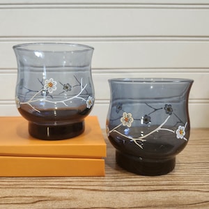 May include: Two gray glass tumblers with a floral design. The tumblers have a dark base and a wider top. The floral design features white branches with small flowers in white, orange, and black. The tumblers are on an orange box.