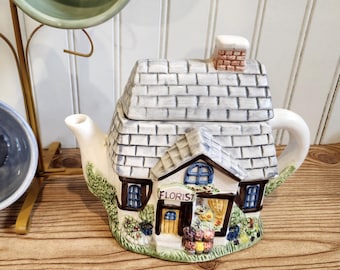 May include: A novelty ceramic teapot in the shape of a house, with a white tiled roof and the word "FLORIST" on the front. A blue teacup and a green bowl are on a gold-coloured metal stand. The items are on a wooden surface.