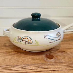 May include: Cream ceramic soup tureen with a dark green lid and handle. The tureen features a rooster and mushroom design in blue, brown, and green. The tureen is on a wooden surface.