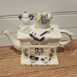 May include: A white ceramic teapot with a house-like design, featuring a small teapot and a basket of fruit on top. The teapot has gold accents and blue details. The handle and spout are curved.