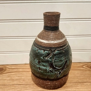 May include: A ceramic vase with a narrow neck and a rounded body. The upper portion features horizontal brown and white bands, while the lower section is textured with a deep teal glaze over a dark brown base, showing carved details.