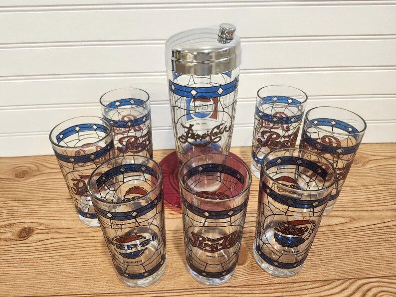 May include: A vintage Pepsi-Cola cocktail set featuring a chrome-topped shaker and six matching highball glasses. The glasses and shaker have a stained-glass design with blue and red accents, and the Pepsi logo. The set is displayed on a wooden surface.