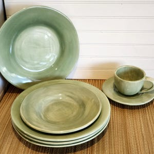 May include: A set of sage green ceramic dinnerware featuring a large bowl, three stacked plates, and a matching teacup on a saucer. Each piece has a smooth, glossy finish with subtle variations in color and a rounded, organic shape.
