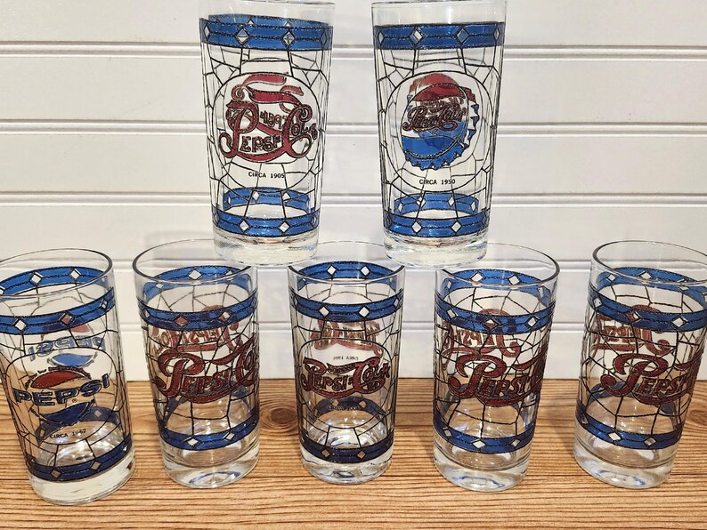 May include: A set of seven vintage Pepsi Cola glasses. Each glass features a stained-glass style design with the Pepsi logo in red, white, and blue. The glasses have a blue border and the text "Circa 1950".