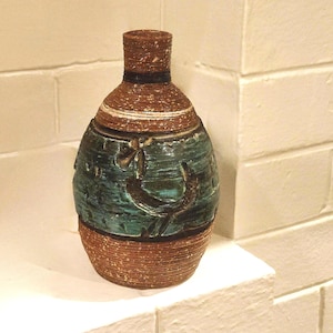 May include: A decorative ceramic vase with a textured brown base and neck, a teal-colored midsection with carved designs, and a dark band. The vase is displayed on a white surface against a white brick wall.