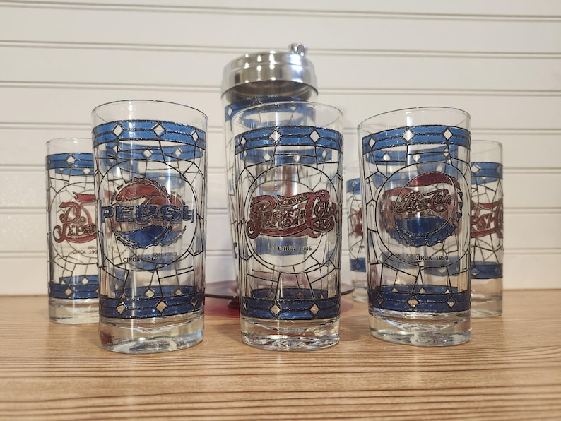 May include: Set of six vintage Pepsi Cola glasses with a blue and white stained glass design. Each glass features the Pepsi logo in red and blue, with the text "Chicago 1902". A stainless steel cocktail shaker is in the background.