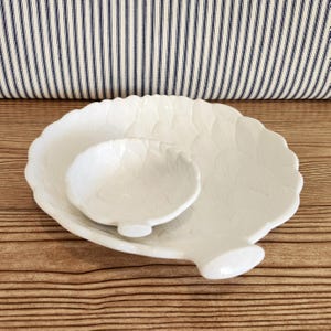 May include: A white ceramic serving set featuring a large, leaf-shaped platter and a smaller, matching bowl. The set has a textured surface with a leaf pattern. The platter has a handle-like extension. The background includes a striped pillow.