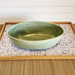 May include: An oval-shaped ceramic bowl with a smooth, glossy finish in a soft sage green color. The bowl features subtle variations in tone across its surface, with a deep basin and gently curved, rounded edges that create a simple, open form.