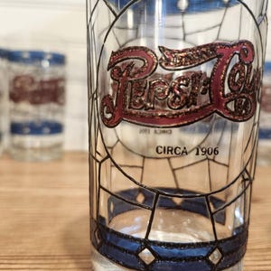 May include: Clear glass Pepsi-Cola drinking glass with a stained glass design. The Pepsi logo is in red and gold, with the text "CIRCA 1906" below. Other glasses are visible in the background.