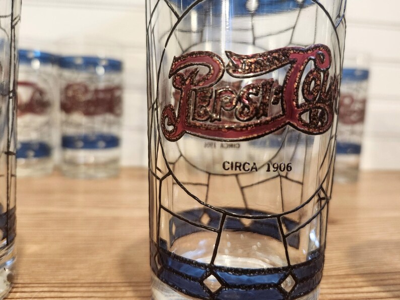 May include: Clear glass Pepsi-Cola drinking glass with a stained glass design. The Pepsi logo is in red and gold, with the text "CIRCA 1906" below. Other glasses are visible in the background.