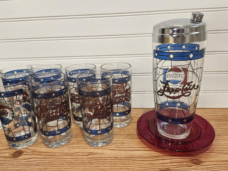 May include: A vintage Pepsi-Cola cocktail shaker and glasses set. The clear glasses and shaker feature a stained-glass design with blue and red accents. The set includes a shaker, a coaster, and six glasses.