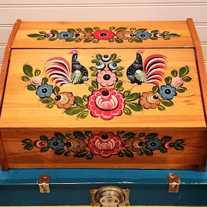 May include: Wooden breadbox with a hinged lid, featuring hand-painted folk art. The design includes two roosters, floral patterns in red, blue, and pink, and green leaves. The box is made of light-colored wood.