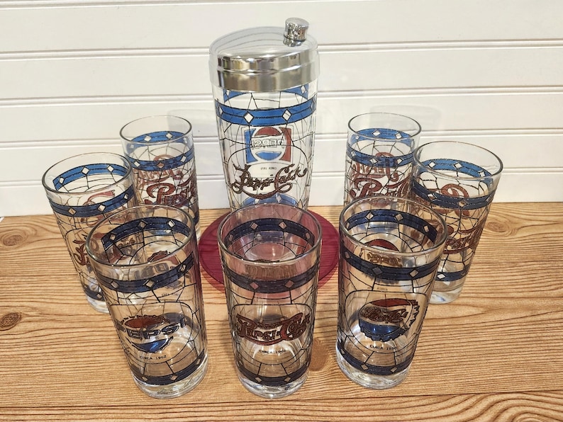 May include: A vintage Pepsi-Cola cocktail shaker and glasses set. The clear glasses and shaker feature a stained-glass design with the Pepsi logo in red, white, and blue. The set includes a metal-topped shaker and six glasses.
