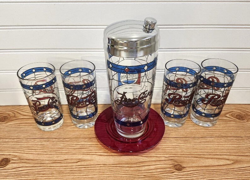 May include: A vintage Pepsi-Cola cocktail set featuring a chrome-topped shaker, a red glass coaster, and four matching highball glasses. The glasses and shaker have a stained-glass design with blue and red accents, and the Pepsi logo.