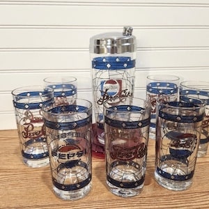 May include: A vintage Pepsi-Cola cocktail set featuring a cocktail shaker and six highball glasses. The glasses and shaker have a stained-glass design with blue and white accents and the Pepsi logo. The set is on a wooden surface.