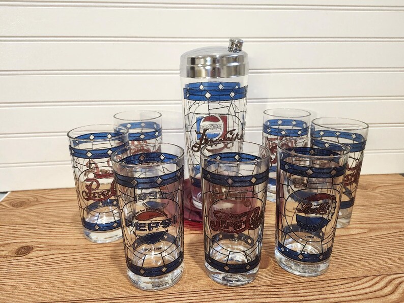 May include: A vintage Pepsi-Cola cocktail set featuring a cocktail shaker and six highball glasses. The glasses and shaker have a stained-glass design with blue and white accents and the Pepsi logo. The set is on a wooden surface.