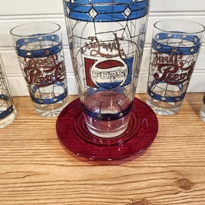 May include: A vintage Pepsi-Cola glass set featuring a large glass, a red plate, and four smaller glasses. The glasses have a stained-glass design with blue and white accents and the Pepsi logo.