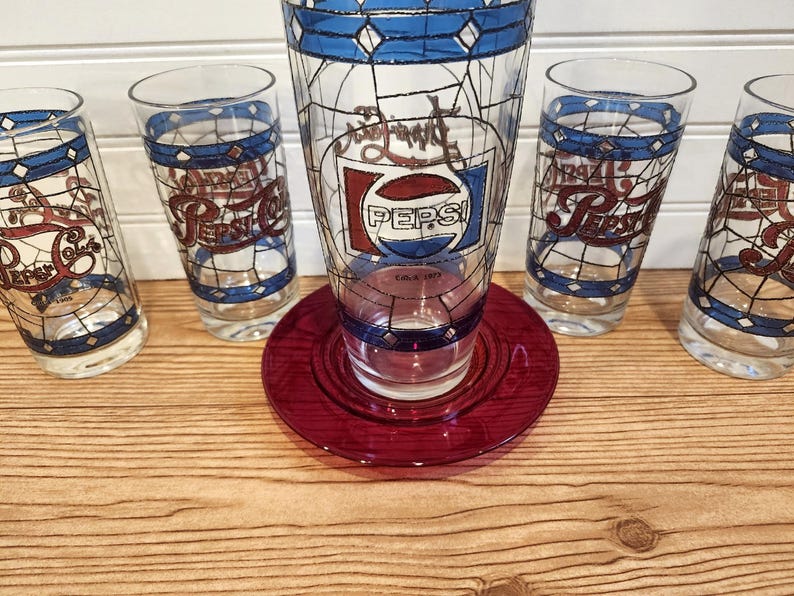 May include: A vintage Pepsi-Cola glass set featuring a large glass, a red plate, and four smaller glasses. The glasses have a stained-glass design with blue and white accents and the Pepsi logo.