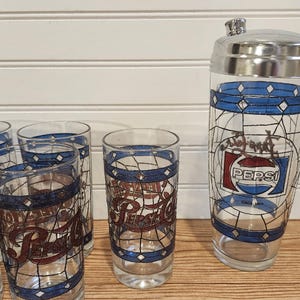 May include: A vintage Pepsi-Cola cocktail shaker and glasses set. The clear glass pieces feature a blue and white geometric design with the Pepsi logo in red and white. The shaker has a silver lid.