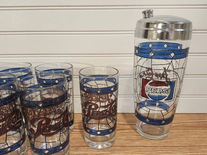 May include: A vintage Pepsi-Cola cocktail shaker and glasses set. The clear glass pieces feature a blue and white geometric design with the Pepsi logo in red and white. The shaker has a silver lid.