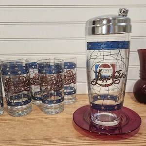 May include: A vintage Pepsi-Cola cocktail shaker and glasses set. The shaker has a silver lid and is on a red plate. The glasses and shaker feature a blue and red design with the Pepsi logo.