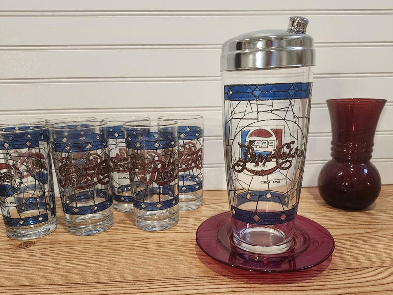 May include: A vintage Pepsi-Cola cocktail shaker and glasses set. The shaker has a silver lid and is on a red plate. The glasses and shaker feature a blue and red design with the Pepsi logo.