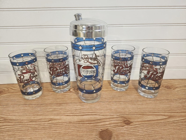 May include: A vintage Pepsi-Cola cocktail shaker set. The set includes a tall shaker with a silver lid and four matching glasses. Each piece features a blue and brown stained-glass design with the Pepsi logo.