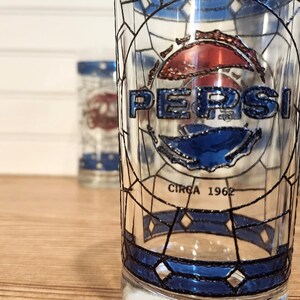 May include: Clear glass Pepsi glasses with a stained glass design. The Pepsi logo is in blue and red, with the text "CIRCA 1962" below. The glasses have a blue border and are on a wooden surface.