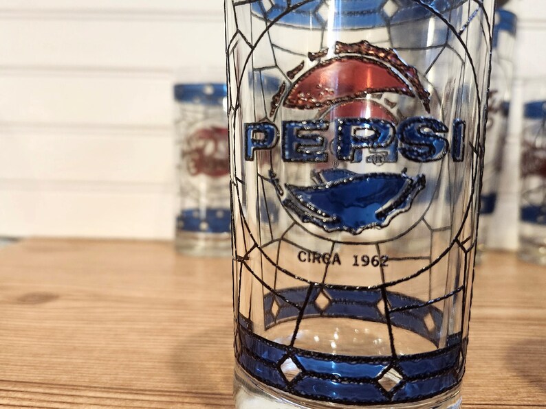 May include: Clear glass Pepsi glasses with a stained glass design. The Pepsi logo is in blue and red, with the text "CIRCA 1962" below. The glasses have a blue border and are on a wooden surface.