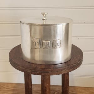 May include: A silver tea canister with a lid and a small handle on top. The word "TEA" is embossed on the front. The canister sits on a small, round, wooden stool with three legs.