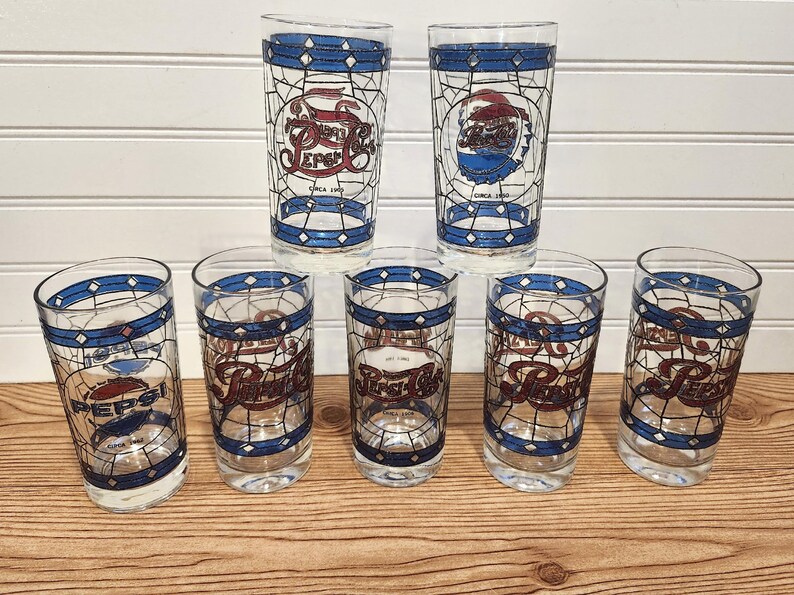 May include: Set of seven vintage Pepsi cola glasses. Each glass features a stained-glass style design with the Pepsi logo in red, white, and blue. The glasses have a blue band around the rim and base, with the words "Circa 1990".
