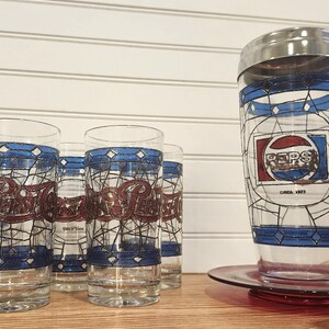May include: A collection of vintage Pepsi glasses and a cocktail shaker. The glasses feature the Pepsi logo in red and blue, with a stained-glass design. The shaker has a silver lid and a similar design, with "PEPS" and "Circa 1923" printed on it.