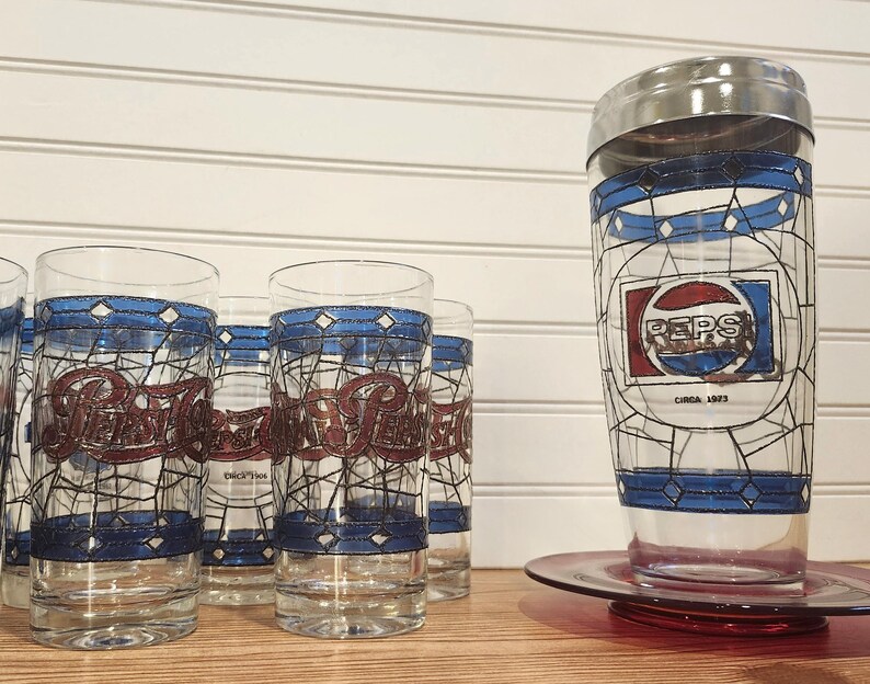 May include: A collection of vintage Pepsi glasses and a cocktail shaker. The glasses feature the Pepsi logo in red and blue, with a stained-glass design. The shaker has a silver lid and a similar design, with "PEPS" and "Circa 1923" printed on it.