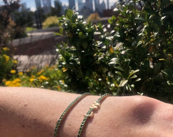 May include: Two bracelets on a person's wrist. One bracelet is a thin gold chain with green stones. The other bracelet is a gold letter 'W' with green stones.