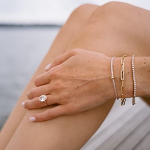 May include: A woman's hand wearing a large, clear, oval-shaped ring with a gold band. The hand is wearing three bracelets on the wrist: a thin, light-colored string bracelet, a gold chain bracelet with oval links, and a diamond tennis bracelet.