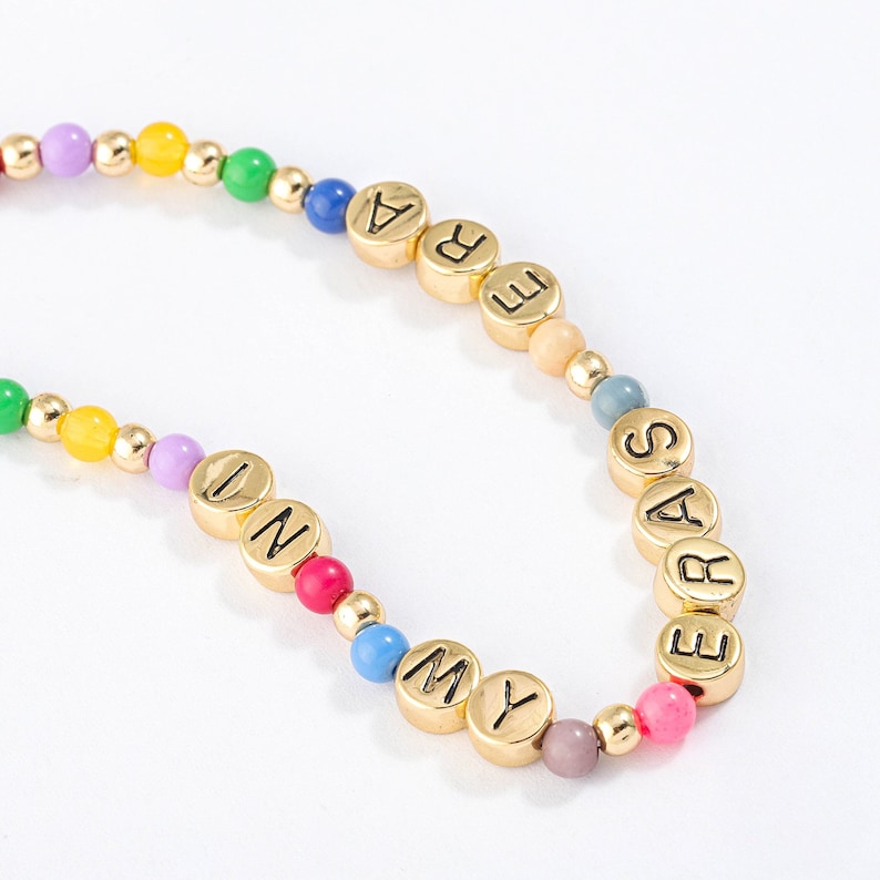 May include: A colorful beaded bracelet with gold letter beads spelling out "ERAS MY UN", with a rainbow color scheme.